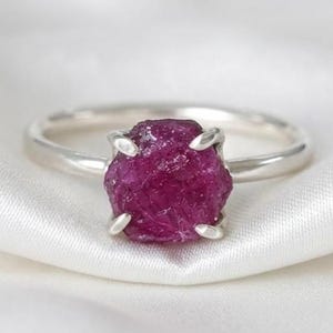 以下が含まれることがあります： 4本の金属製の爪で留められた、未加工でカットされていない深いマゼンタ色の宝石を特徴とするシルバーリング。リングのバンド部分は滑らかで磨かれており、中央の石の質感のある不規則な表面と対照的です。