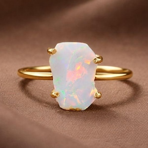 May include: A gold ring featuring a large, irregular-shaped opal gemstone. The opal is primarily white with flashes of iridescent color. The ring's band is a simple, polished gold.