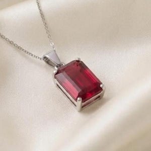 May include: A silver-toned necklace featuring a rectangular, emerald-cut deep red gemstone pendant held in a four-prong setting. The pendant is suspended from a delicate, thin silver-toned metal chain that connects to a triangular bail.