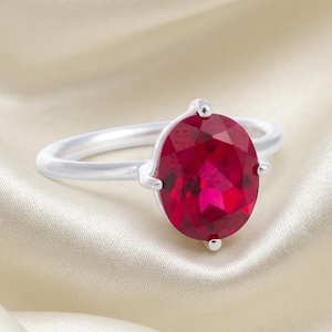 May include: A silver ring featuring a large, oval-cut, vibrant red gemstone held in place by four prongs. The band is smooth and polished, showcasing a clean, metallic finish that complements the deep red color of the central stone.