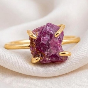 May include: A gold ring featuring a single, raw, deep magenta-colored gemstone set in a four-prong setting. The stone has an irregular, faceted, and textured surface, while the band is smooth, polished, and thin, creating a contrast between the metal.