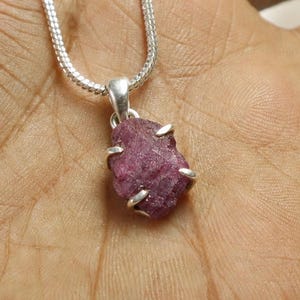 May include: A pendant necklace featuring a raw, deep pink gemstone set in a silver-toned metal prong setting. The pendant hangs from a thin, flexible silver-toned chain. The stone has an irregular, textured surface and a faceted appearance.