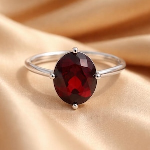 May include: A silver ring with a large, oval-shaped, deep red gemstone. The gemstone is the central feature, set in a simple band with small decorative details either side. The ring is shown on a gold-toned fabric.