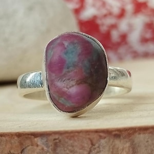 May include: A silver ring with a large, oval stone in shades of pink and teal. The stone is set in a polished silver band. The ring is presented on a wooden surface, showcasing the contrasting colours and textures of the materials.