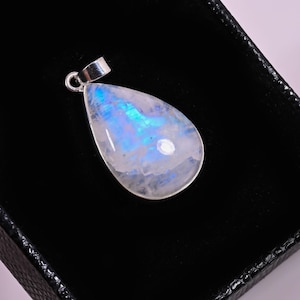 May include: A pear-shaped moonstone pendant with a silver bail. The gemstone displays a milky white base with iridescent blue and purple flashes. The pendant is set against a black background.