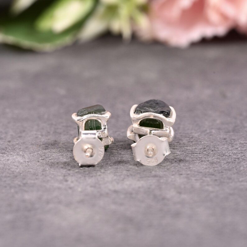 May include: A pair of sterling silver stud earrings. Each earring features a rough-cut, dark green gemstone set in a silver bezel. The earrings are displayed on a gray surface, with a blurred floral background.