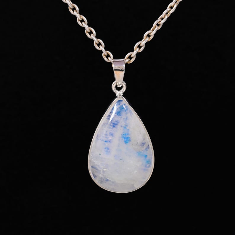 May include: A silver chain necklace with a teardrop-shaped pendant. The pendant features a polished, pearlescent moonstone with blue and white hues. The necklace is displayed against a black background.