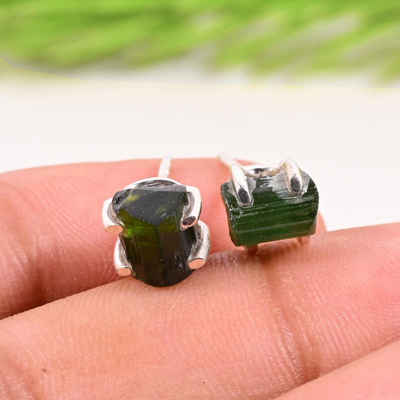 May include: A pair of stud earrings featuring raw, dark green tourmaline gemstones set in silver. The gemstones have a natural, rough texture and are mounted on simple silver posts, showcasing the unique beauty of the raw crystals.