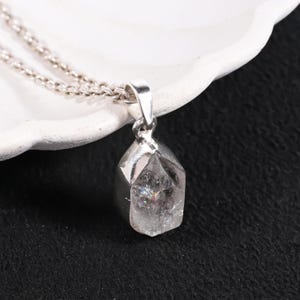 Pu&ograve; includere: Una collana con pendente in argento con un cristallo trasparente e sfaccettato. Il cristallo presenta inclusioni interne a forma di arcobaleno ed &egrave; sospeso a una delicata catena d'argento. Il pendente &egrave; posto su una superficie bianca e uno sfondo nero.