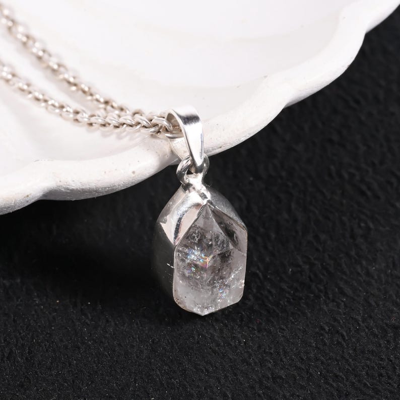 Pu&ograve; includere: Una collana con pendente in argento con un cristallo trasparente e sfaccettato. Il cristallo presenta inclusioni interne a forma di arcobaleno ed &egrave; sospeso a una delicata catena d'argento. Il pendente &egrave; posto su una superficie bianca e uno sfondo nero.