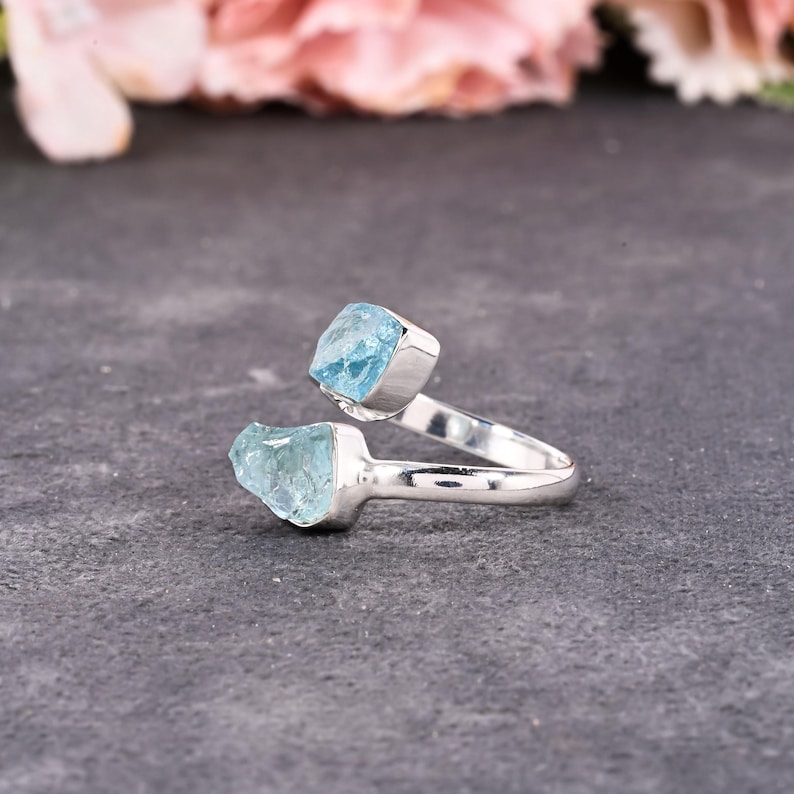 May include: A silver ring featuring two raw, light blue aquamarine gemstones. The ring's band curves around the finger, with each stone set in a square bezel. The gemstones have a natural, rough texture.