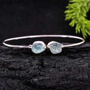 May include: A silver-toned bangle bracelet featuring two light blue, raw-cut aquamarine stones. The stones are set on either end of the thin, curved band. The bracelet is displayed on a dark, textured surface, with a green plant in the background.