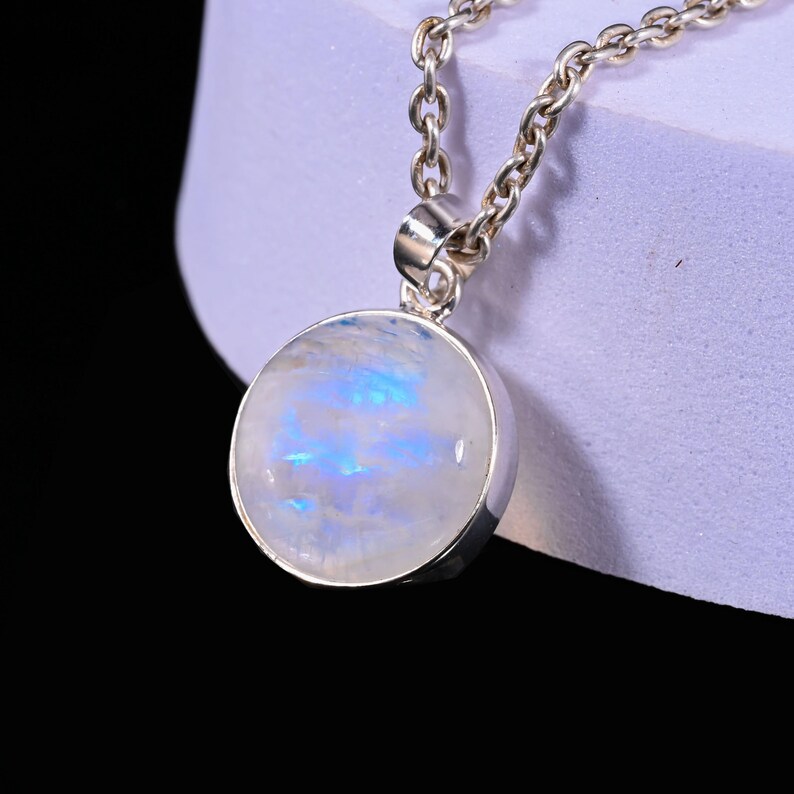 May include: A silver necklace featuring a round pendant with a moonstone. The moonstone has a white base with blue and iridescent highlights. The chain is a classic link style.