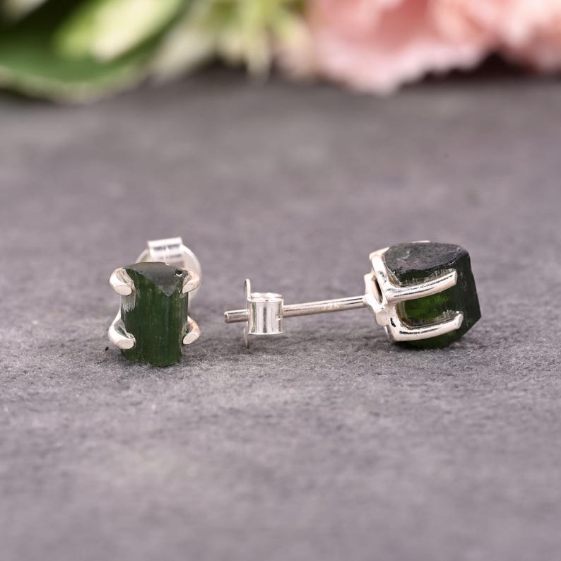 May include: A pair of stud earrings featuring raw, dark green tourmaline gemstones set in silver. The gemstones have a natural, unpolished appearance, and the silver settings have a simple, claw-like design. The earrings are displayed on a gray surface.