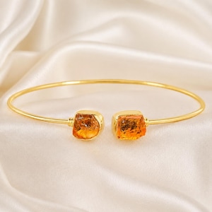 Könnte beinhalten: Ein goldfarbenes Armreif-Armband mit zwei quadratischen, bernsteinfarbenen Edelsteinen. Das Armband ist vor einem weißen, seidigen Hintergrund platziert, der das Design und die Farbe des Schmucks hervorhebt.