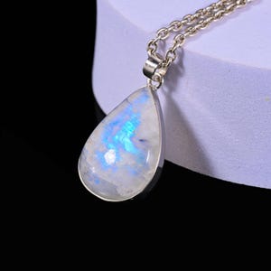 May include: A teardrop-shaped pendant necklace with a silver chain. The pendant features a large, iridescent moonstone with blue and white hues. The stone is set in a silver bezel.