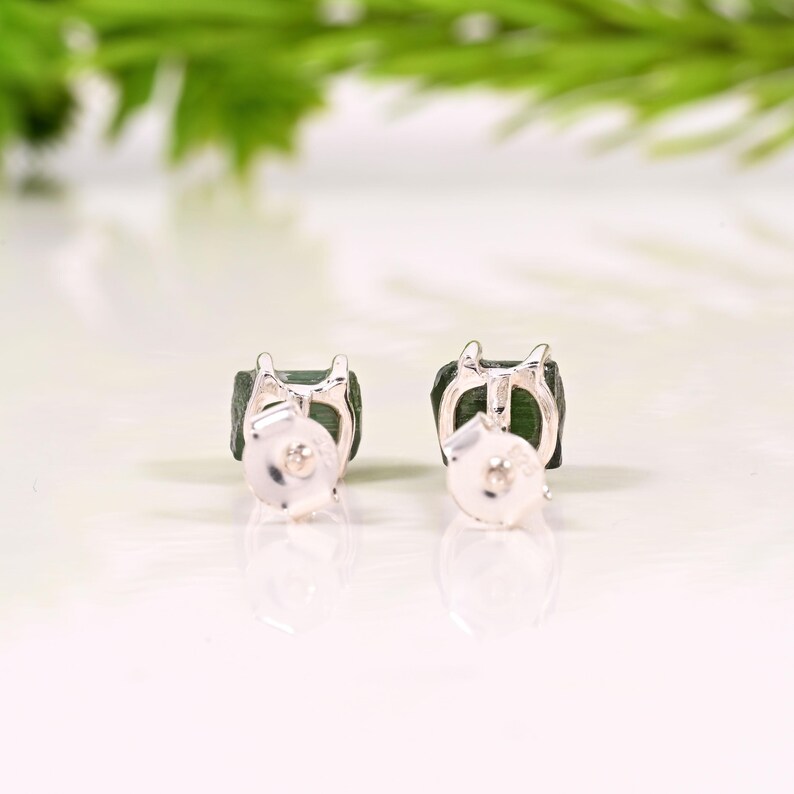 May include: A pair of square-cut, dark green gemstone stud earrings with silver metal settings and backs. The earrings are displayed on a white surface with a blurred green background.