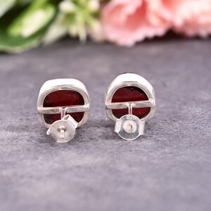 Pode incluir: Um par de brincos de bot&atilde;o quadrados com uma pedra preciosa vermelha escura emoldurada em metal prateado. Os brincos t&ecirc;m um fecho de pino e borboleta. O fundo &eacute; uma mistura desfocada de cinza e rosa.
