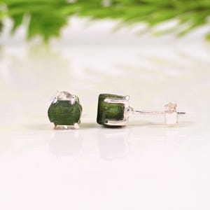 May include: A pair of stud earrings featuring raw, dark green tourmaline gemstones set in silver. The gemstones have a natural, textured appearance. The earrings are displayed on a white surface with a blurred green background.