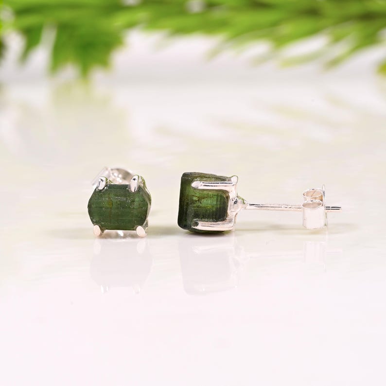 May include: A pair of stud earrings featuring raw, dark green tourmaline gemstones set in silver. The gemstones have a natural, textured appearance. The earrings are displayed on a white surface with a blurred green background.