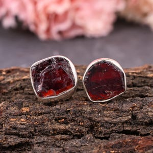 Pode incluir: Dois brincos de bot&atilde;o prateados com pedras de granada vermelha escura bruta. Os brincos est&atilde;o colocados sobre uma superf&iacute;cie de madeira castanha texturizada, com um fundo floral rosa desfocado.