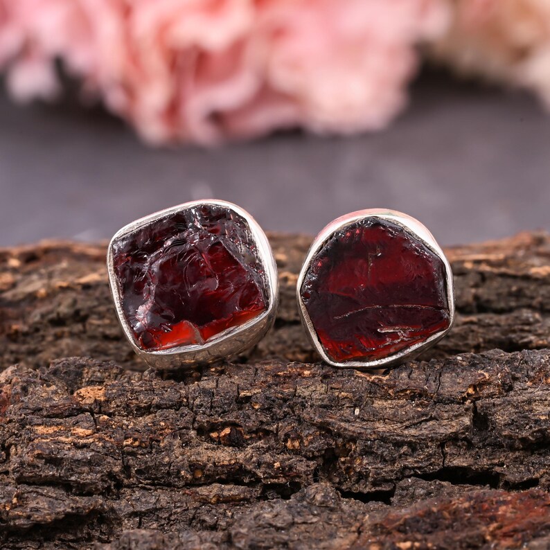 Pode incluir: Dois brincos de bot&atilde;o prateados com pedras de granada vermelha escura bruta. Os brincos est&atilde;o colocados sobre uma superf&iacute;cie de madeira castanha texturizada, com um fundo floral rosa desfocado.