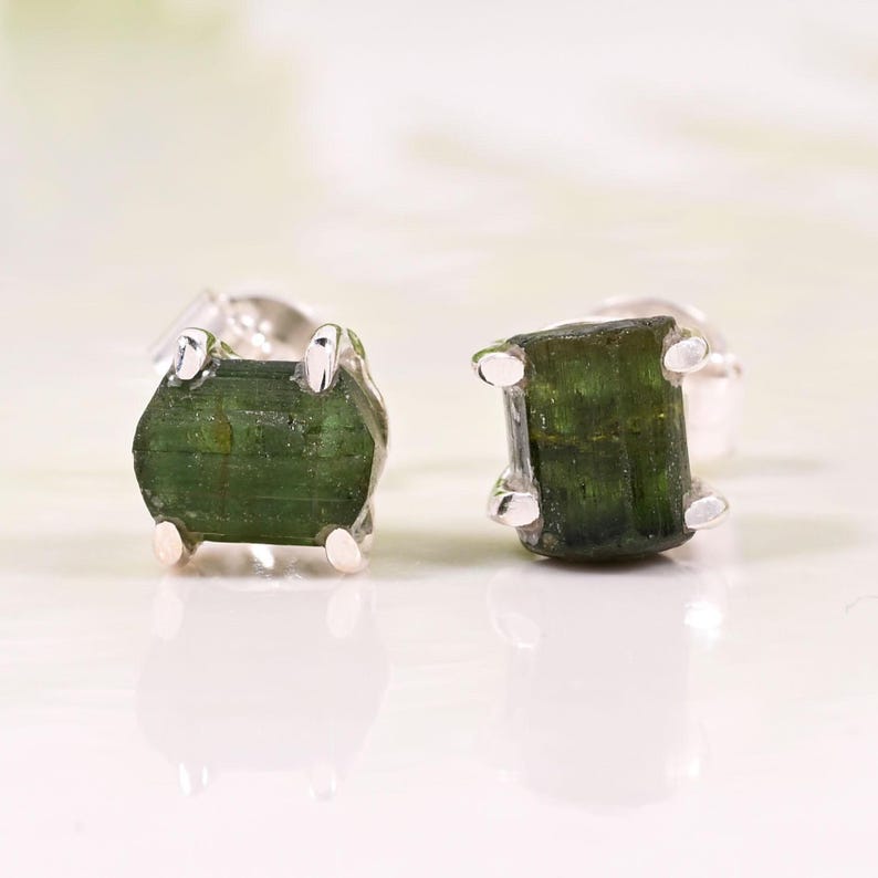 May include: A pair of stud earrings featuring raw, rectangular green tourmaline gemstones set in silver. The gemstones have a natural, textured appearance and are mounted with four silver prongs. The earrings are displayed on a white surface.