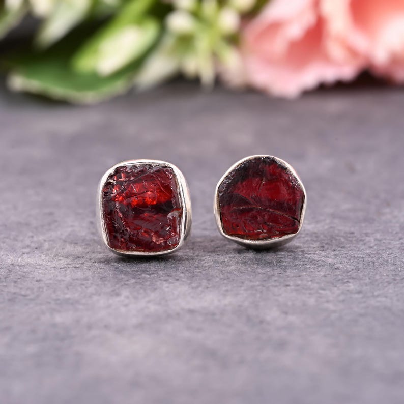 Pode incluir: Um par de brincos quadrados. Cada brinco apresenta uma pedra de granada vermelha escura bruta, embutida em uma moldura prateada. As granadas t&ecirc;m uma superf&iacute;cie &aacute;spera e texturizada. O fundo &eacute; cinza desfocado.