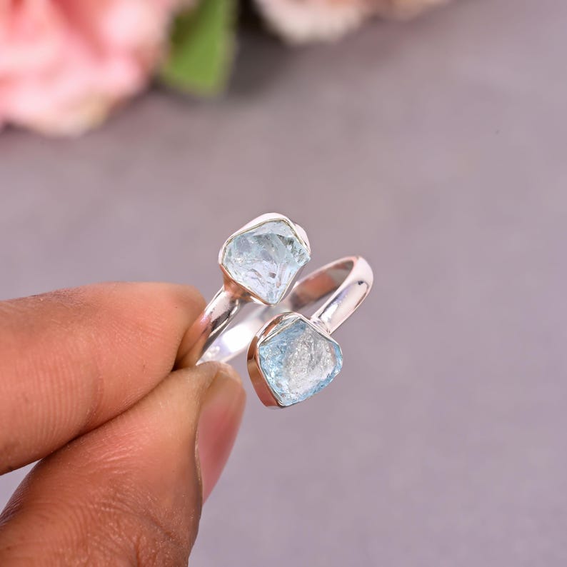 May include: A silver ring featuring two light blue, irregularly shaped gemstones. The ring's band curves around the finger, with one stone at the top and the other at the bottom. The gemstones have a rough, natural appearance.