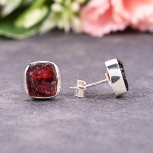 Pode incluir: Um par de brincos de bot&atilde;o quadrados. Cada brinco apresenta uma pedra de granada vermelha escura bruta embutida em uma moldura de prata. Os brincos s&atilde;o exibidos em uma superf&iacute;cie cinza, com um fundo floral desfocado.