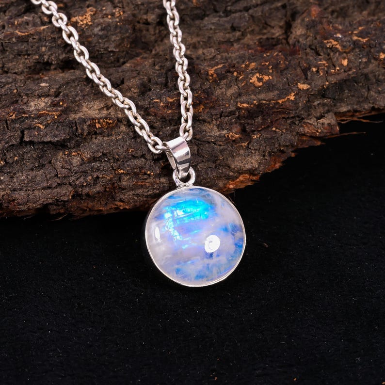 May include: A silver necklace featuring a round, iridescent moonstone pendant. The moonstone displays a mix of blue and white hues, set in a silver bezel. The chain is a classic link style, creating a simple yet elegant piece of jewelry.