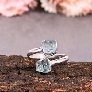 May include: Two sterling silver rings with light blue, raw-cut gemstones. The rings are displayed on a dark, textured surface, with a blurred floral background. The gemstones have a natural, irregular shape.