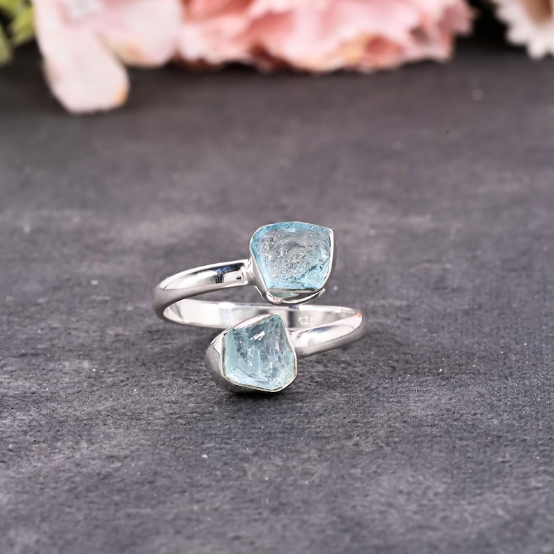 May include: A silver ring featuring two raw, light blue aquamarine stones. The ring's band curves around the finger, with one stone at the top and the other at the bottom. The stones have a natural, unpolished appearance, set against a dark gray background.