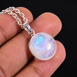 May include: A round, iridescent moonstone pendant necklace. The moonstone is set in a silver bezel and hangs from a delicate silver chain. The moonstone displays a blue and white color. The necklace is shown against a dark background.