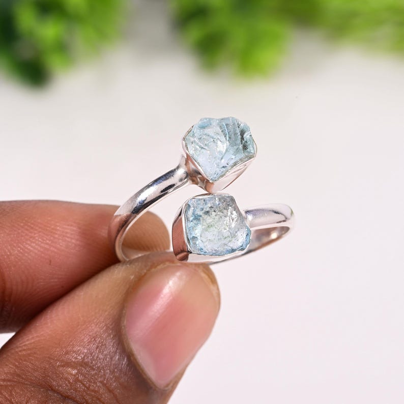 May include: A silver ring featuring two raw, light blue aquamarine gemstones. The ring's band curves around the finger, with each stone set in a bezel setting. The gemstones have a natural, rough texture.