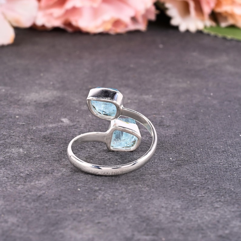 May include: A silver ring with two light blue gemstones. The ring's band curves around the finger, with the gemstones set in square bezels. The ring is displayed on a dark gray surface, with pink flowers in the background.