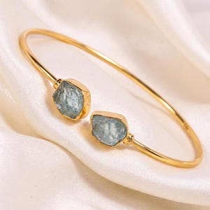 Könnte beinhalten: Ein goldfarbenes, offenes Armband mit zwei rauen, hellblauen Aquamarinsteinen an den Enden. Das Metallband ist dünn und kreisförmig, wobei die Gravur 14K auf der Innenseite des Bandes in der Nähe einer der Steinfassungen sichtbar ist.
