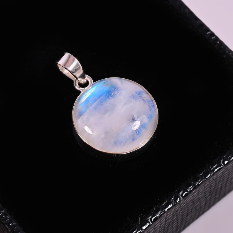 May include: A round moonstone pendant with a silver bail. The moonstone has a milky white base with blue and iridescent highlights. The pendant is set in a silver bezel and is displayed against a black background.