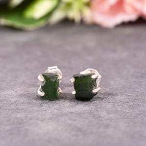 May include: A pair of dark green tourmaline stud earrings. Each earring features a raw, rectangular-shaped green tourmaline stone held in place by silver prongs. The earrings are set against a gray background, with soft floral elements in the background.