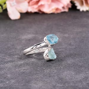 May include: A silver-toned ring featuring two raw, light blue aquamarine gemstones. The ring's band curves around the finger, with one stone at the top and the other at the bottom. The gemstones have a rough, natural texture.