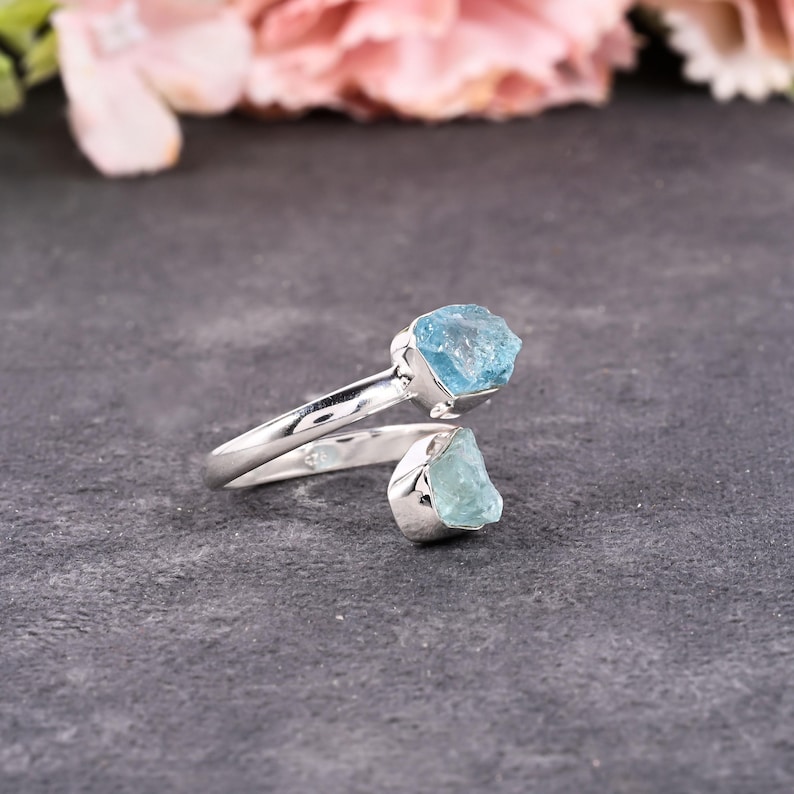 May include: A silver-toned ring featuring two raw, light blue aquamarine gemstones. The ring's band curves around the finger, with one stone at the top and the other at the bottom. The gemstones have a rough, natural texture.