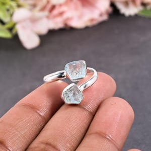 May include: A silver ring featuring two rough-cut, light blue aquamarine gemstones. The ring's band curves around the finger, with the stones set at opposite ends. The background is a blurred mix of pink and gray.