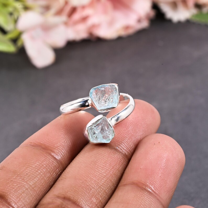 May include: A silver ring featuring two rough-cut, light blue aquamarine gemstones. The ring's band curves around the finger, with the stones set at opposite ends. The background is a blurred mix of pink and gray.