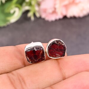 Pode incluir: Um par de brincos de bot&atilde;o quadrados. Cada brinco apresenta uma pedra de granada vermelha escura bruta embutida em uma moldura de prata. Os brincos s&atilde;o exibidos em uma m&atilde;o, com um fundo desfocado de verde e rosa.
