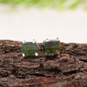 May include: A pair of raw green tourmaline stud earrings. Each earring features a rough-cut, rectangular green tourmaline stone set in a silver metal base. The earrings are displayed on a textured, brown wooden surface.