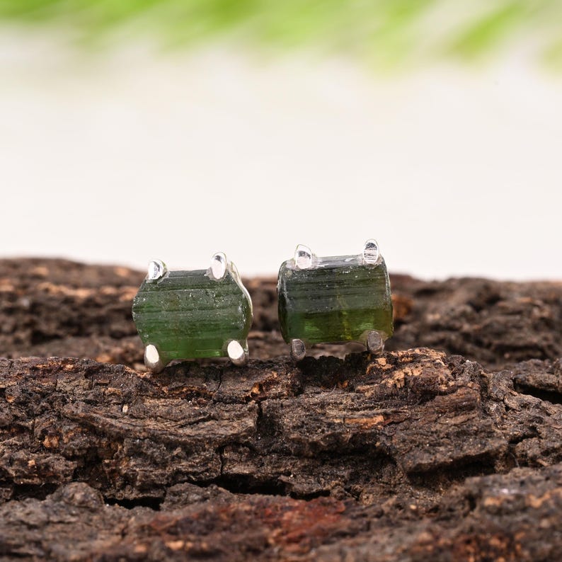 May include: A pair of raw green tourmaline stud earrings. Each earring features a rough-cut, rectangular green tourmaline stone set in a silver metal base. The earrings are displayed on a textured, brown wooden surface.