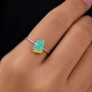 以下が含まれることがあります： 鮮やかな原石オパールをあしらったシルバーリング。オパールは青、緑、黄色など様々な色合いを見せています。リングは細いシルバーのバンドにセットされており、石の自然な美しさを際立たせています。