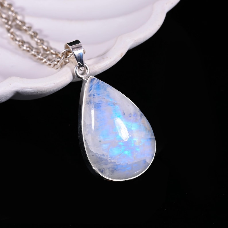 May include: A teardrop-shaped pendant necklace featuring a large, iridescent moonstone. The moonstone has a pearlescent, blue and white glow, set in a silver-colored metal. The chain is also silver-colored and delicate. The necklace is displayed against a white background.