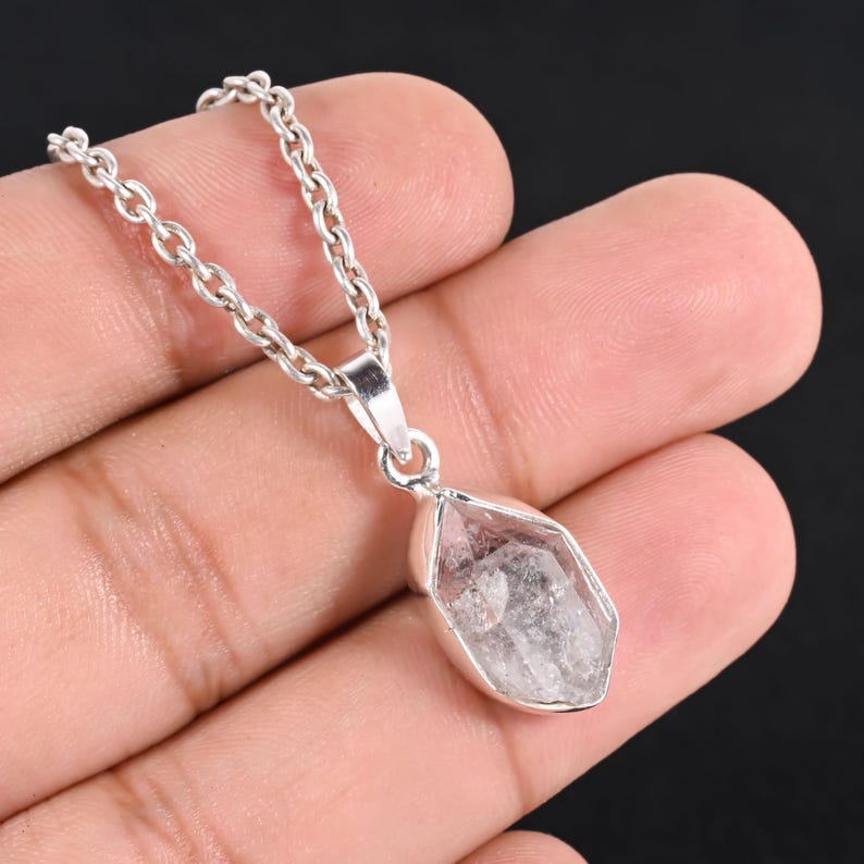 Pu&ograve; includere: Una collana con pendente in cristallo di quarzo trasparente con una catena d'argento. Il cristallo di quarzo &egrave; sfaccettato e incastonato in una lunetta d'argento. La collana &egrave; tenuta in mano su sfondo scuro.