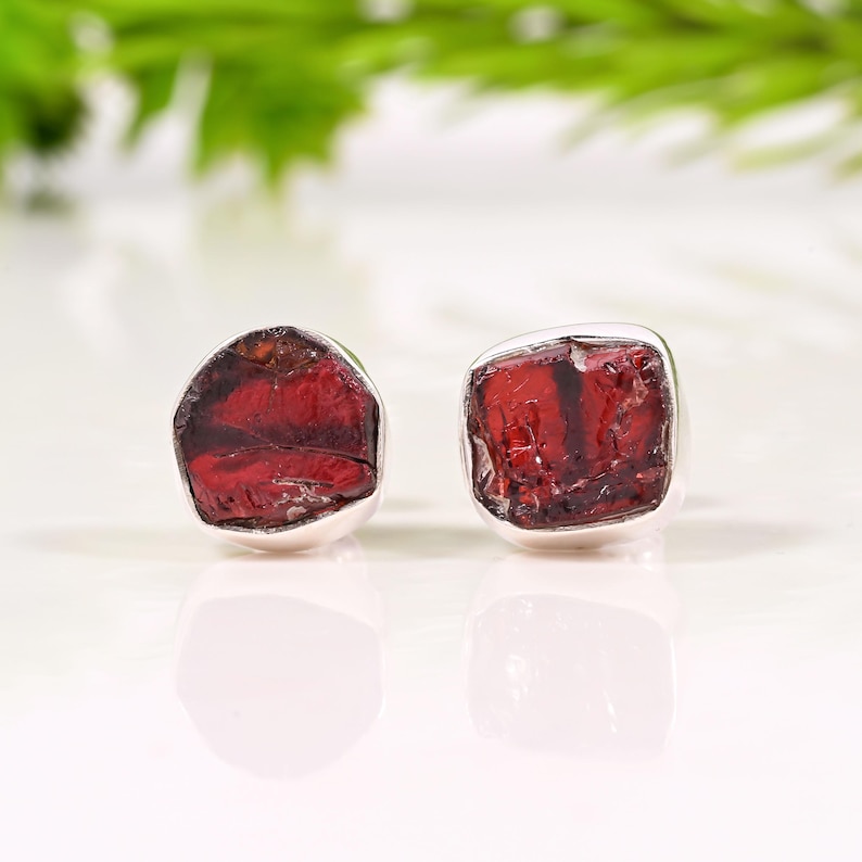 Pode incluir: Um par de brincos de press&atilde;o com pedras de granada vermelha escura bruta, emolduradas em prata. As pedras preciosas t&ecirc;m uma textura natural &aacute;spera. Exibido em um fundo branco com folhagem verde.